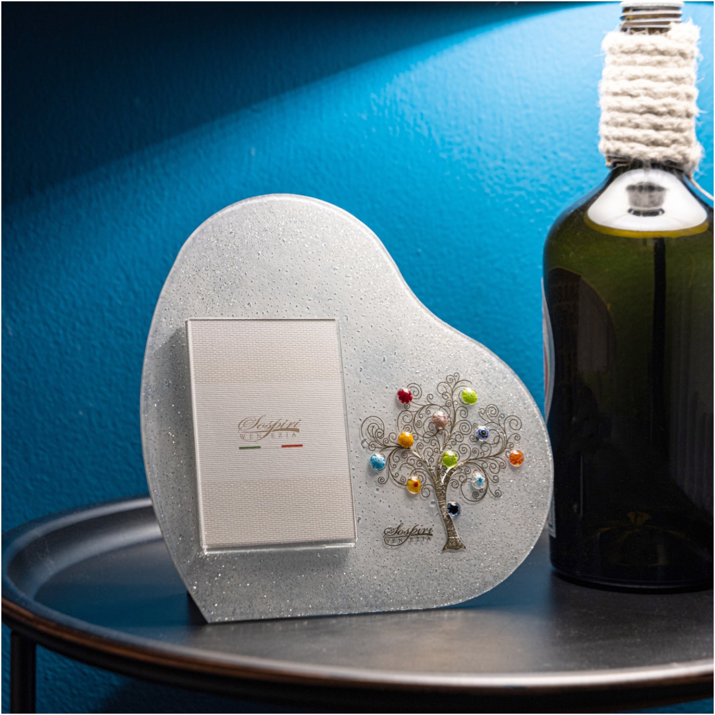 Cornice portafoto bianca a forma di cuore in vetro di Murano con decoro Albero della Vita in oro zecchino e murrine di Murano colorate, appoggiata su un tavolino nero con sfondo blu.